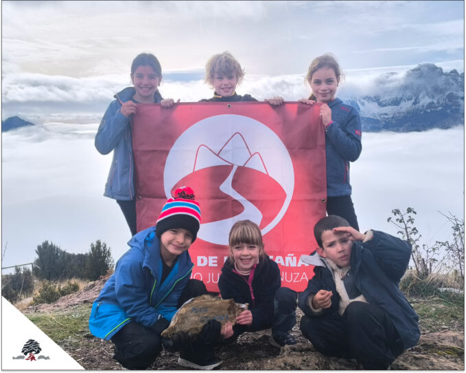 Grupo de niños y jóvenes del Club de Montaña del Colegio Juan de Lanuza en Panticosa, posando con una bandera en un paisaje nevado del Pirineo.