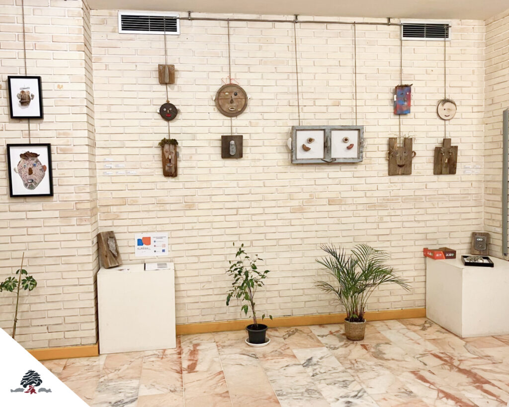 Vista de la exposición “Caras y Caretas” en el Colegio Juan de Lanuza, con esculturas y máscaras elaboradas con distintos materiales, expuestas sobre una pared de ladrillo claro.